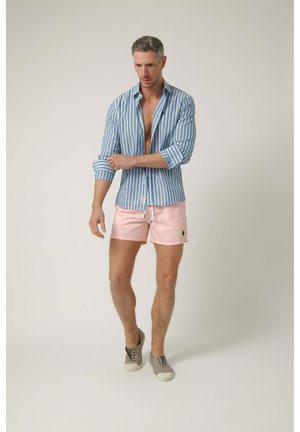 Hombre remangando las mangas de una camisa a rayas azul y blanca, con pantalones cortos rosa claro y zapatos grises sin cordones, de pie frente a un fondo liso.