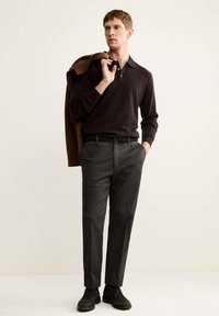Polo shirt scuro e pantaloni scuri su misura. Il modello tiene una giacca marrone sulle spalle; scarpe nere con finitura testurizzata. Sfondo neutro.