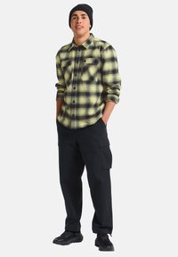 Chemise en flanelle à carreaux jaunes et noirs, dotée de deux poches poitrine. Associée à un pantalon cargo noir et des baskets, avec un bonnet noir.