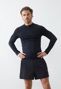 Črn dolg rokav športni top z četrtim zadrgo in rebrasto teksturo, v kombinaciji s črnimi kratkimi hlačami. Obe stvari imata eleganten, prilegajoč se dizajn.