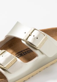 Silverfärgade metalliska Birkenstock-sandaler med två justerbara remmar, som har en beige korkfotbädd och en texturerad gummisula. Guldtonade spännen.