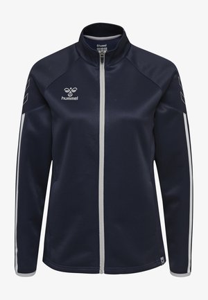 Marineblauwe ritsjack met witte accentstrepen, gemaakt van gladde stof. Heeft een opstaande kraag en het 'hummel' logo op de bovenste linkerkant.