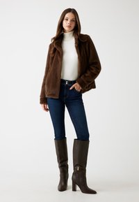 Giacca di pelliccia sintetica marrone con colletto, abbinata a un dolcevita bianco, jeans blu scuro e stivali al ginocchio in pelle marrone con zip dorata.
