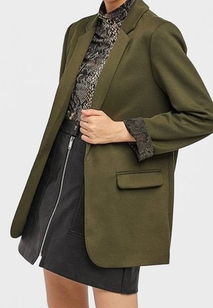 Kvinde iført en olivengrøn blazer over en slangeprintbluse og en sort lædernederdel med lynlås, med den ene hånd holdende på blazerlappen.