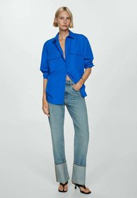 Camicia blu con bottoni e due taschini sul petto, abbinata a jeans di denim chiaro con fondo svasato. Il modello indossa sandali neri.