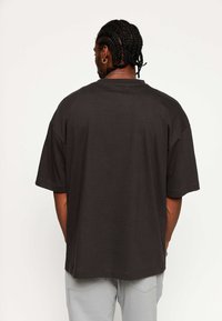 Hombre con cabello trenzado que lleva una camiseta negra holgada y pantalones gris claro, de pie de espaldas a la cámara frente a un fondo neutro.