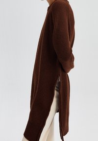 Brauner Strickpullover mit langen Ärmeln, lockerer Passform und seitlichen Reißverschlüssen für anpassbare Länge. Strukturierter Stoff mit geripptem Muster.