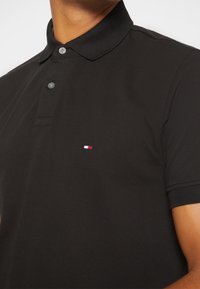 Polo shirt en coton noir avec un tissu texturé, deux boutons au col et un petit logo rouge, blanc et bleu sur la poitrine.