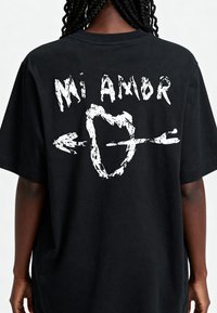 T-shirt noir avec un texte graphique en blanc, en gras "Mi Amor" accompagné d'un design de flèche et de cœur au dos. Matière en coton doux, coupe décontractée.