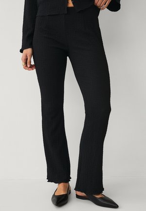 Kvinde iført teksturerede sorte højtaljede bukser kombineret med sorte slingback-flade sko med spidst tåparti mod en ensfarvet baggrund.