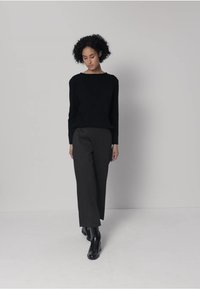 Musta neulepusero, jossa pyöreä pääntie, yhdistetty harmaisiin leveälahkeisiin housuihin. Malli on pukeutunut mustiin nilkkureihin, taustana neutraali värimaailma.