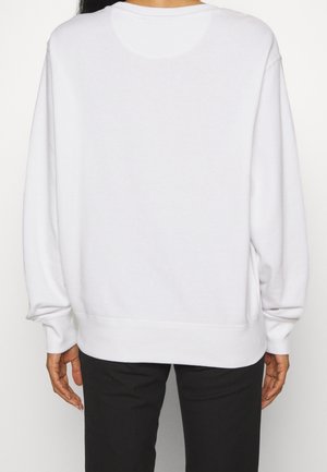 Osoba oblečená v jednoduché bílé mikině s dlouhým rukávem a černých kalhotách, zobrazená zezadu na neutrálním pozadí.