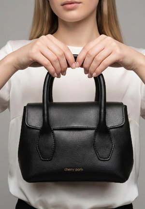 Femme portant une chemise blanche tenant un sac à main en cuir noir avec des doubles poignées et un logo doré "cherry paris" sur le devant.