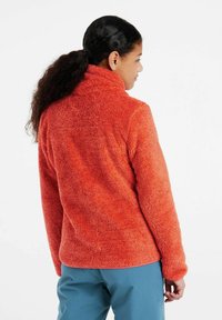 Plyšová oranžová fleece bunda s vysokým golierom a dlhými rukávmi, kombinovaná s modrými nohavicami. Tkanina má mäkký, textúrovaný vzhľad.