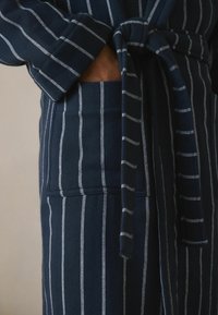 Jūras spēku svītrains mēteli, kas izgatavots no mīksta auduma, ar saitēm viduklī, garām piedurknēm un priekšējo kabatu ar kontrastējošām baltām svītrām.