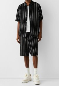 Camicia a maniche corte a righe nere e pantaloni corti abbinati di lunghezza al ginocchio, realizzati in tessuto leggero, abbinati a scarpe da ginnastica bianche e calzini alla caviglia.