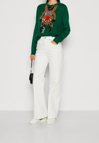 Grüner Strickpullover mit einem braunen Tiergrafik und rotem Pompon, kombiniert mit hoch taillierten weißen weitem Hosen und Sneakers. Schwarze Handtasche.