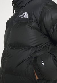 Černá péřová bunda má zapínání na zip vpředu, vysoký límec, elastické manžety a vyšívané logo. Na rukávu je výrazné označení výplňového výkonu 700.