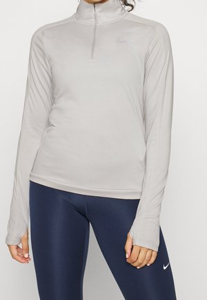 Kobieta ubrana w jasnoszarą, sportową bluzę z długim rękawem i ćwiartkowym zamkiem oraz granatowe legginsy Nike, stojąca na jednolitym tle.