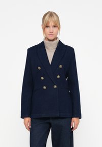 Marinblå dobbeltspent blazer med gullknapper, to framlommer og en skreddersydd design. Brukes over en lys beige høyhalset genser.