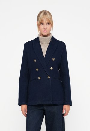 Blazer bleu marine croisé avec des boutons dorés, deux poches avant et une coupe ajustée. Porté par-dessus un col roulé beige clair.