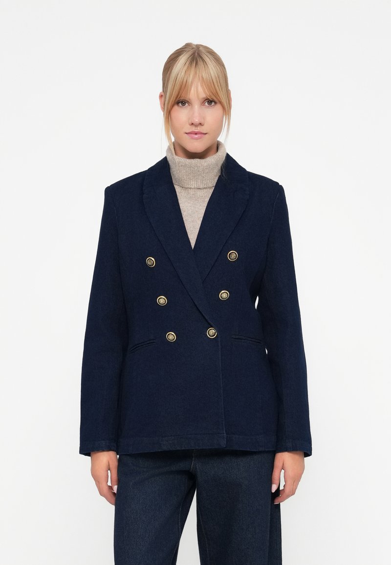 Marinblå dobbeltspent blazer med gullknapper, to framlommer og en skreddersydd design. Brukes over en lys beige høyhalset genser.
