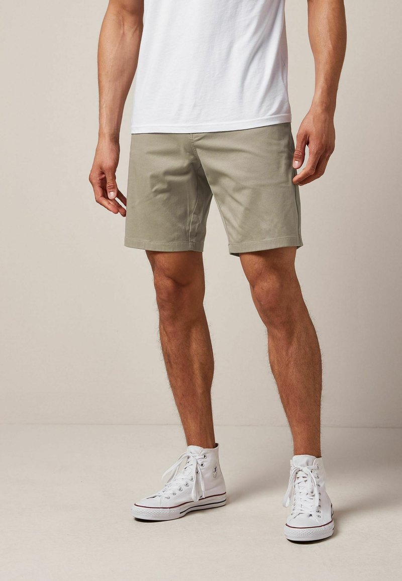 Pantaloncini da uomo fino al ginocchio di colore verde oliva chiaro in un misto cotone liscio. Abbinati a sneakers alte bianche e una t-shirt bianca aderente.