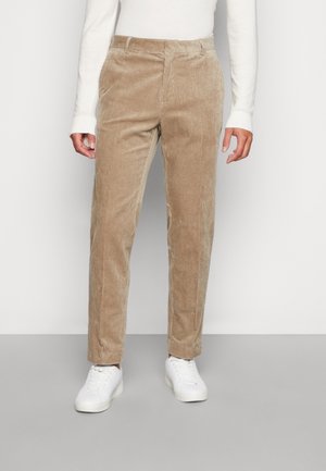 Pantalon beige en velours côtelé coupe droite porté avec un haut blanc à manches longues et des baskets blanches, debout devant un fond uni.