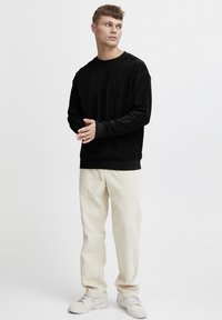 Hanorac negru cu mâneci lungi, guler rotund, textură moale și mansete ribdate. Asortat cu pantaloni bej deschis din catifea și sneakersi albi.