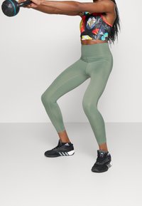 Leggings verdi a vita alta con una texture liscia e cuciture laterali, abbinati a un crop top floreale multicolore e scarpe da ginnastica nere.