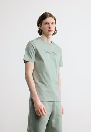 Joven con cabello hasta los hombros que lleva una camiseta Calvin Klein verde claro y pantalones cortos a juego, de pie contra un fondo blanco liso.