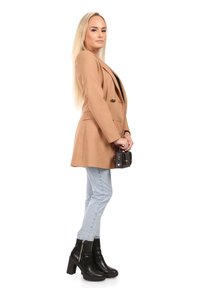 Beige dubbelzijdig colberts, lichtblauwe skinny jeans, zwarte enkellaarsjes met dikke hakken, en een kleine zwarte handtas in de hand.