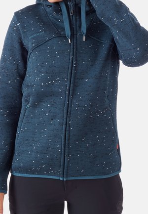 Osoba ubrana w niebieską, nakrapianą bluzę na zamek z sznurkami, stojąca na tle jednolitego białego tła.