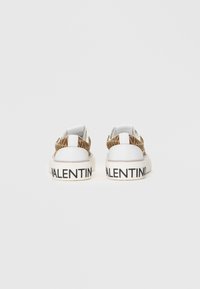 Valge tossud tekstuuriga pruuni seljaga, millel on must "VALENTINO" logo tallal. Kujundatud ümarate vormide ja lõdva stiiliga.