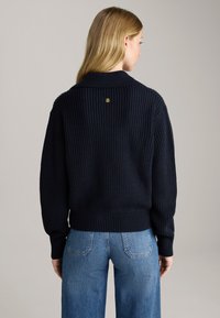 Granatowy sweter z dzianiny o prążkowanej strukturze i złożonym kołnierzem. Posiada małe złote logo z tyłu. W parze z jasnoniebieskimi spodniami o szerokich nogawkach.