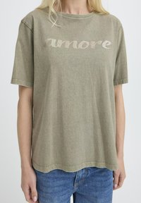 Camiseta de color verde oliva con mangas cortas, que presenta un diseño de texto brilloso que dice "amore". Tela de algodón suave con un corte relajado y dobladillo redondeado.