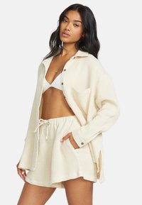 Completo loungewear di colore crema con texture a nido d'ape, composto da una camicia con bottoni dal taglio morbido e pantaloni corti con elastico e cordino, muniti di tasche.