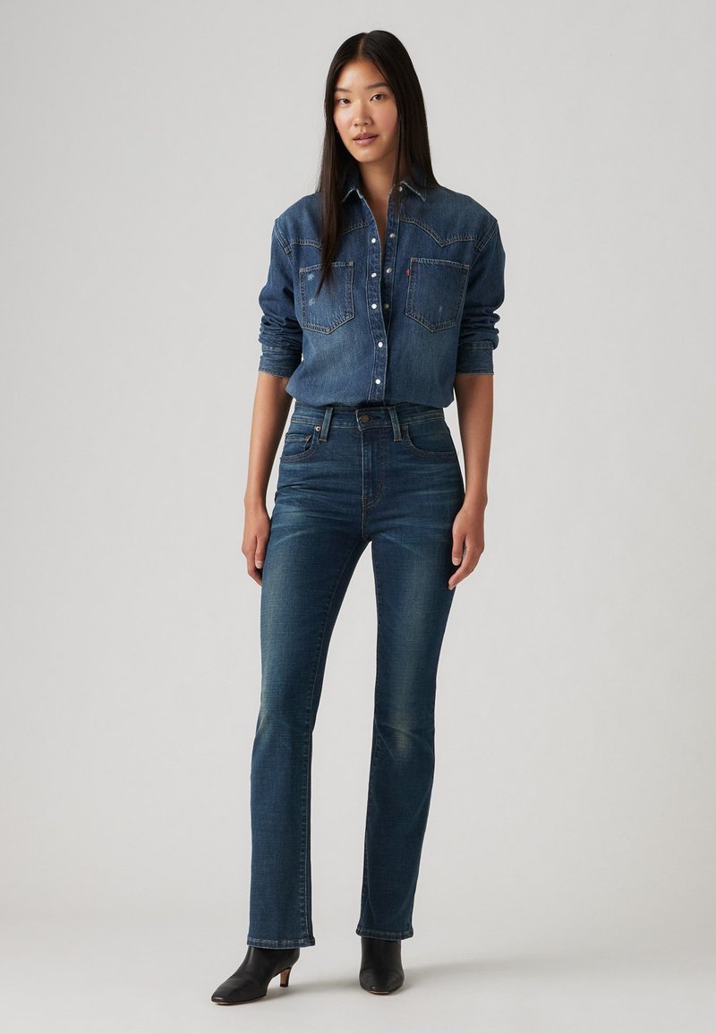 Dunkelblauses Denim-Hemd mit Knopfverschluss und zwei Brusttaschen, kombiniert mit blauen ausgestellten Jeans und schwarzen Stiefeletten mit Absatz. Glatte Textur, taillierte Passform.
