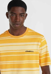 Kollane triibuline T-särk, millel on heledamad kollased jooned, ümmargune kaelus, lühikesed varrukad ja must "ONEILL" logo ees. Puuvillane materjal, lõdva istuvusega.