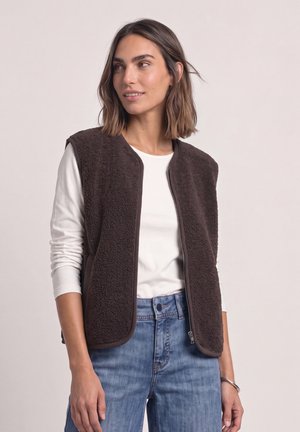 Woman wearing a dark brown fleece vest over a white long-sleeve shirt and blue jeans, looking slightly to the side with a neutral background.