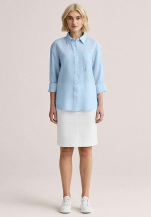 Lyshåret kvinde iført en lyseblå skjorte med knapper og ærmer rullet op, hvid knælængde nederdel og hvide sneakers, stående mod en ensartet baggrund.