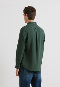 Donkergroen langslijfshirt met een button-down kraag, achterkant met plooi en manchet met witte knopen; gecombineerd met blauwe denim jeans.