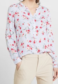 Blus med blommönster i blå och vita ränder, långa ärmar, v-ringad halslinje och knappdetalj. Tyget verkar lätt och mjukt.