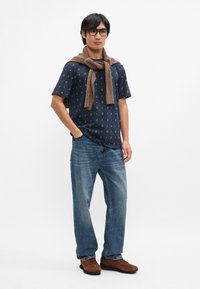 Ναυτικό κοντομάνικο πουκάμισο με λουλουδάτο μοτίβο, φορεμένο με ένα υφασμάτινο καφέ πουλόβερ δεμένο γύρω από το λαιμό, συνδυασμένο με ανοιχτό μπλε τζιν και καφέ παπούτσια.