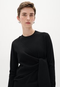 Schwarzer Strickpullover mit langen Ärmeln, der ein gewickeltes Designdetail an der Taille und einen Rundhalsausschnitt aufweist. Weiche Textur und taillierte Silhouette.