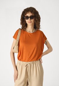 Haut orange à manches courtes avec des accents en dentelle texturée, associé à un pantalon beige à cordon de serrage. Le mannequin porte des lunettes de soleil noires et un collier en perles.