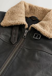 Jachetă din piele maro cu un guler din blană artificială de culoare deschisă. Dispune de un fermoar frontal, nasturi cu cap și o textură netedă cu cusături vizibile.