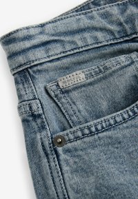 Blugi din denim de culoare albastră deschisă, cu un brâu cusut, buzunar cu închidere prin buton și un etichetă de marcă pe buzunar.