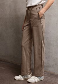 Pantalons beiges ajustés en tissu texturé, dotés d'une taille haute, de poches latérales et d'une silhouette droite, associés à des baskets blanches.