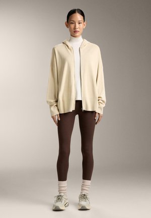 Žena stoji noseći bež jaknu s patentnim zatvaračem, bijelu rolku, smeđe helanke, krem čarape i krem tenisice na jednobojnoj pozadini.